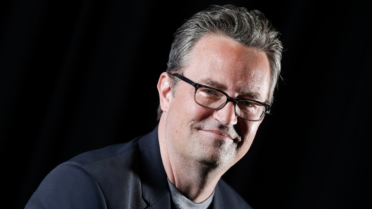 Matthew Perry szemüvegben, sötét háttér előtt mosolyog